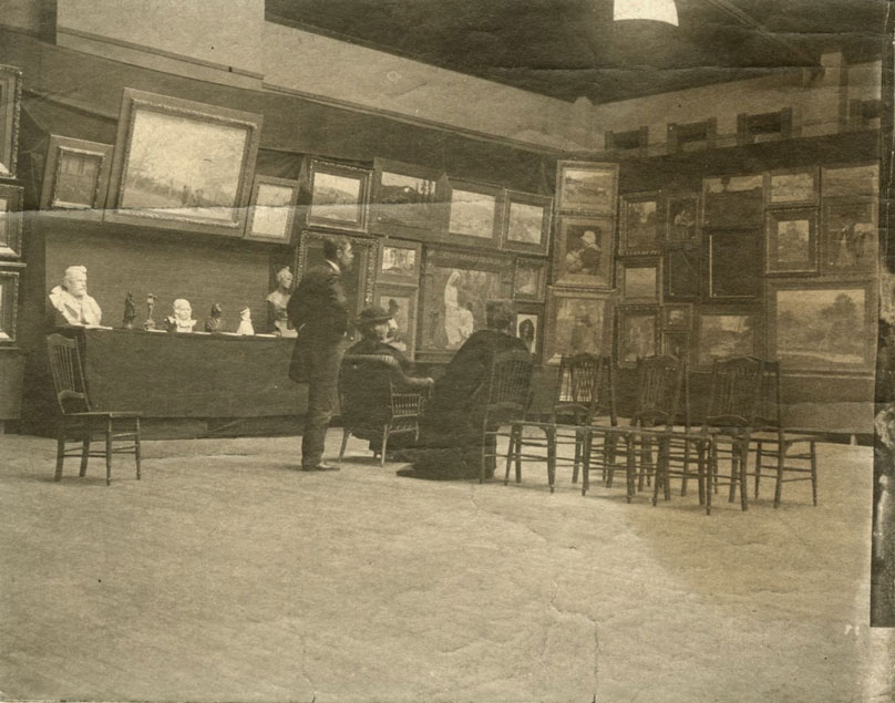 Two women and a man are in the middle of a room with paintings filling the walls and several busts displayed on a table. .