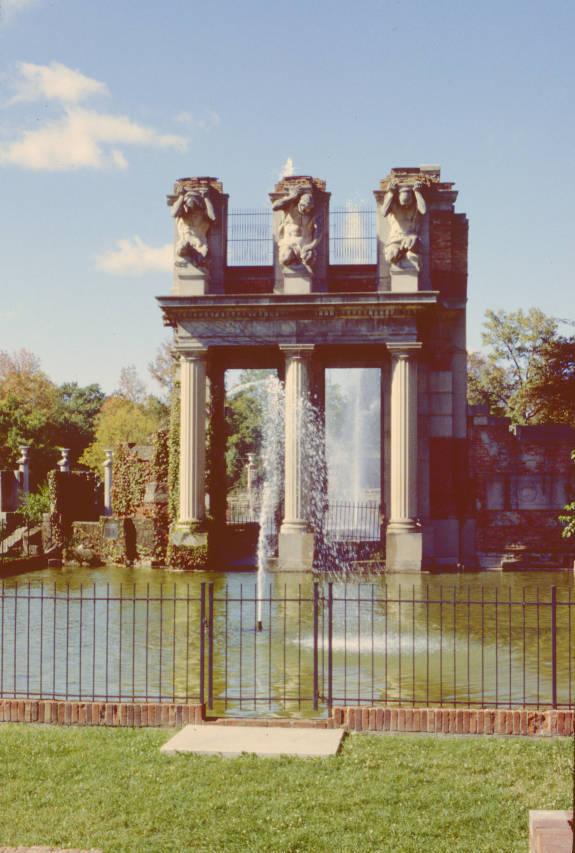 Art piece with three columns and three statues overlooking a pool of water.