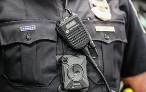 Police officer is shown wearing a body cam in front of a rack of charging body cameras, 2019