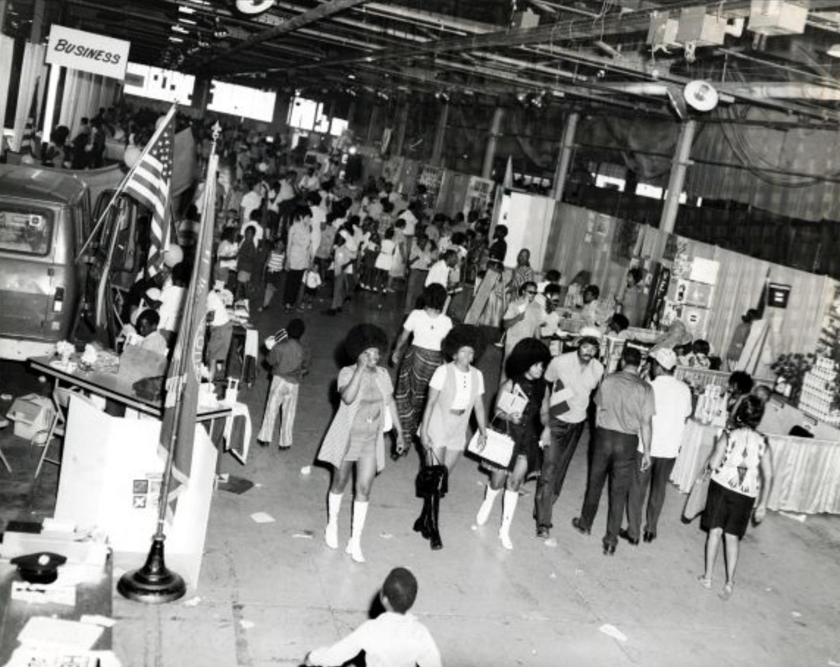 Crowds fill the center aisle in a large building. Display tables and booths line the aisle.