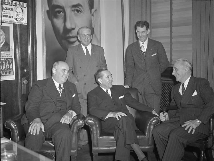 Three men sit in chairs and two other men stand behind them.