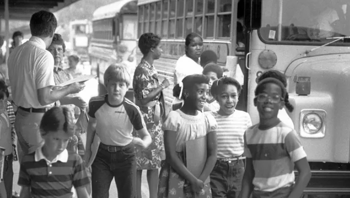 A group of Black and white students are standing outside of a line of busses. A man holding a clipboard and a woman are talking.
