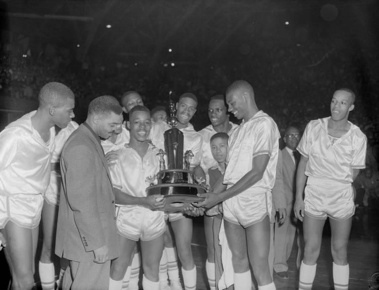 Young men in basketball uniforms stand around a trophy. 