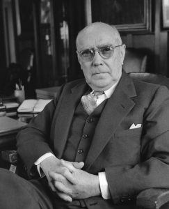 A man in a three-piece suit sits at a desk with his hands folded at his waist.