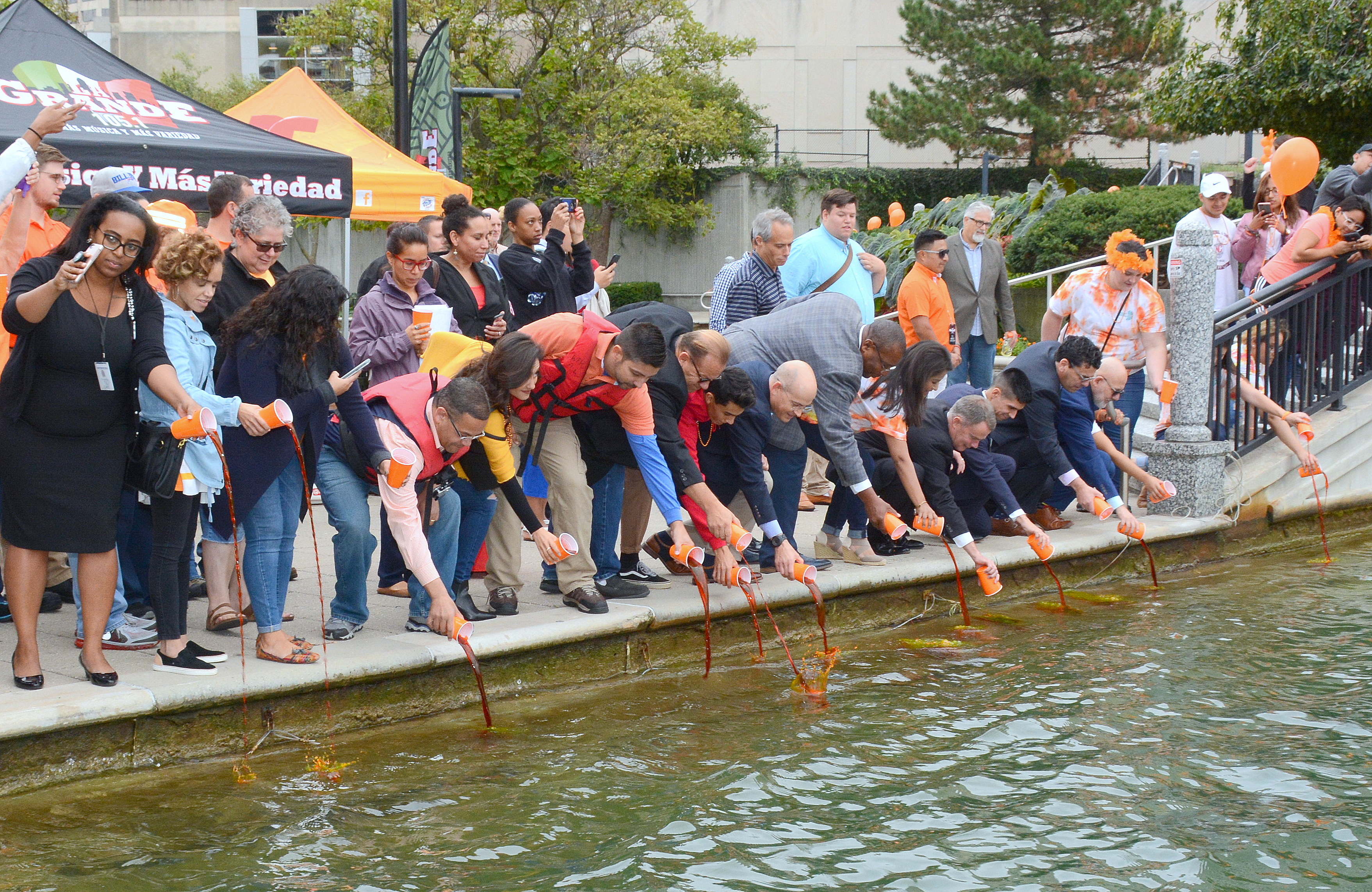 A group of people line the canal. They are all pouring cups of orange dye into it.