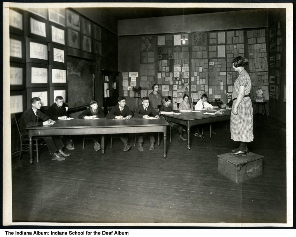 In a classroom, a woman stands on a box in front of a group of students seated at two, large tables.