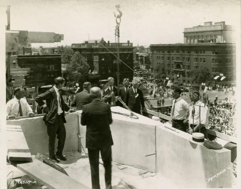 A group of men stand around the newly placed cornerstone of the monument. Crowds of people watch from below.