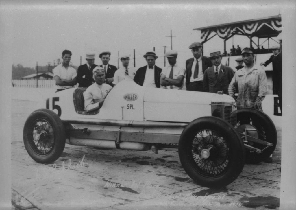 A man sits in an old-fashioned race car. A row of men stand behind the car.