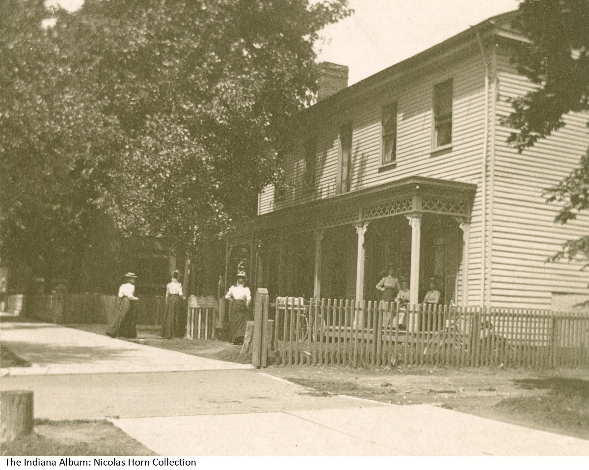 Three women sit on the front porch of a multistory house. Three more women are walking through the fence towards the house.