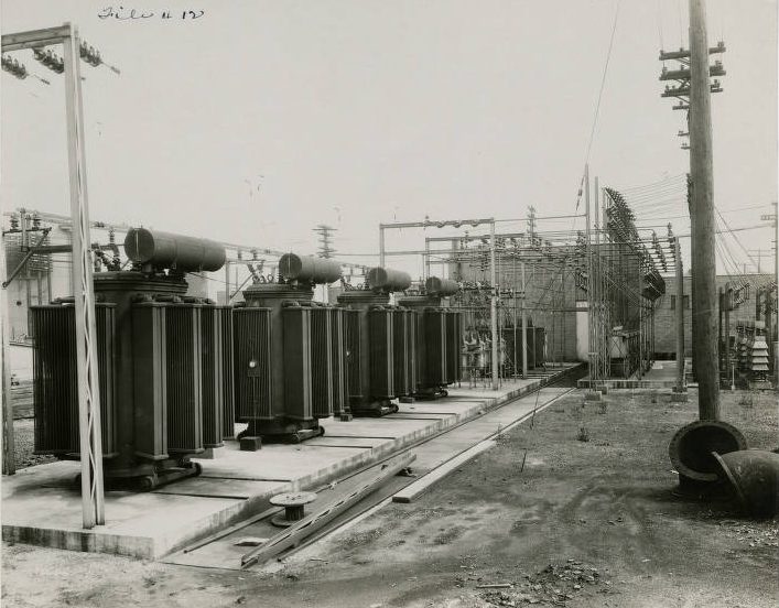 A power station with electric lines throughout. A small brick structure sits in the background and large, round structures in the foreground.