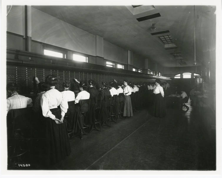 A row of women sit in chairs in front of a long wall of switchboards. Three women stand behind them and supervise. 