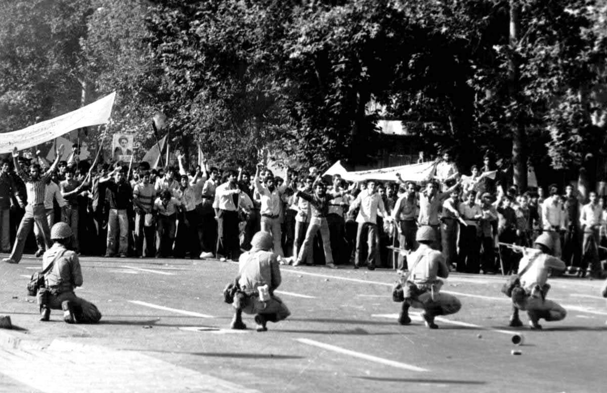 Four armed men are lined up with guns pointed toward a crowd of protestors.