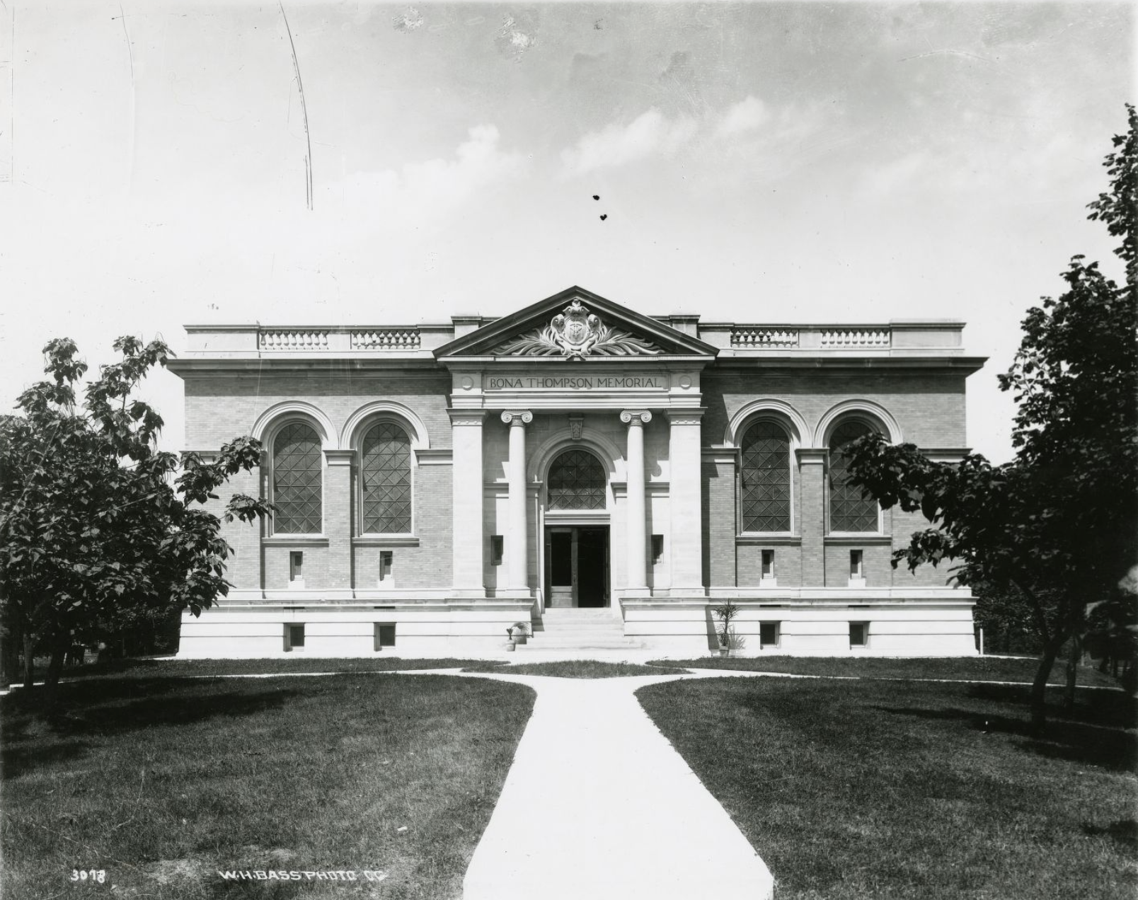 Front view of a two-story Neo-classical building with arched mullioned windows. The roof is flat with a balustrade. The entrance, which extends from the front of the building, is flanked by columns.