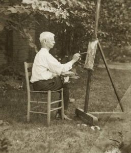 A man is sitting in a chair outside and painting at an easel.