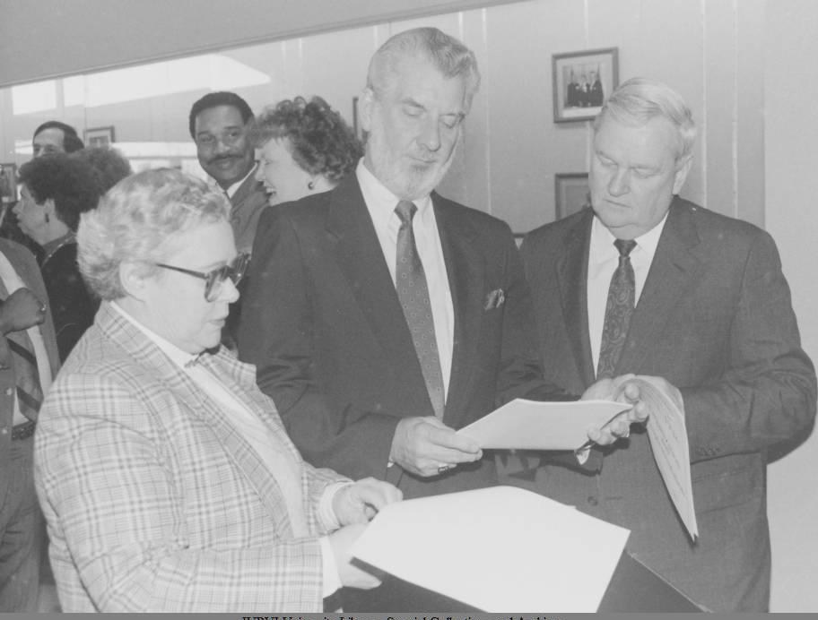 Three people look at a document together. Other people are standing in the background.