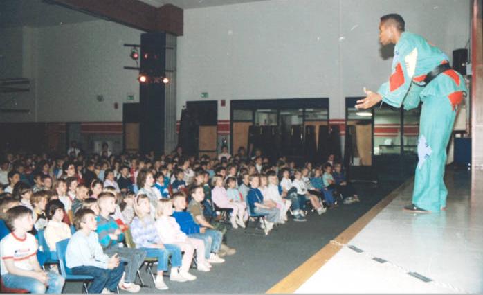 A large audience of children sits in rows in front of a man on a stage.