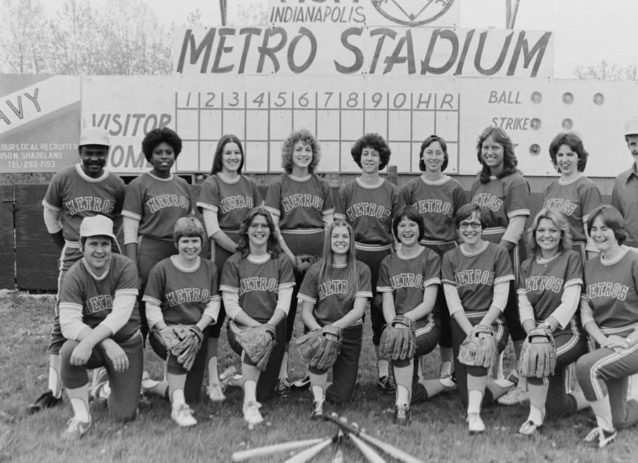 A group of women in softball uniforms pose in front of a scoreboard.