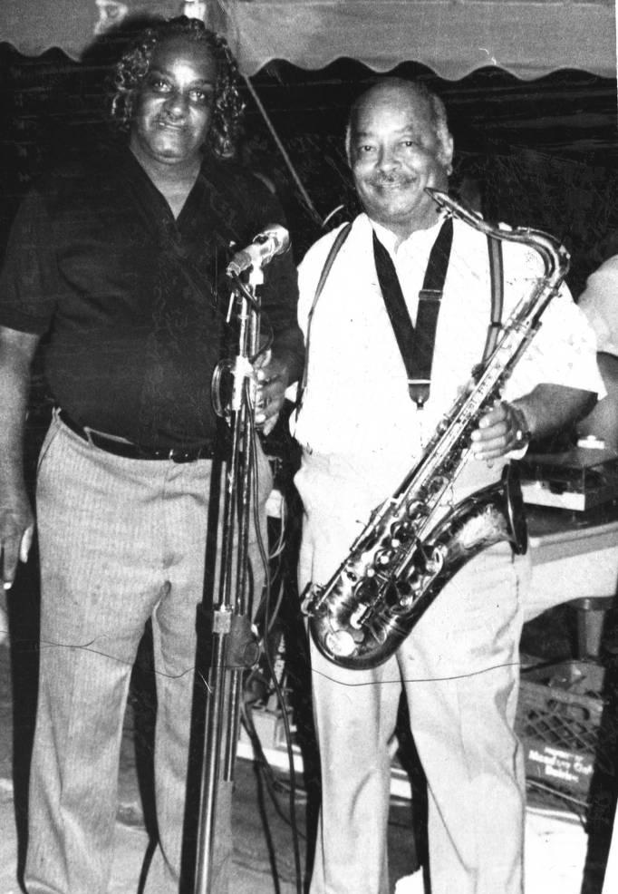 Two men are standing behind a microphone. One is holding a saxophone.