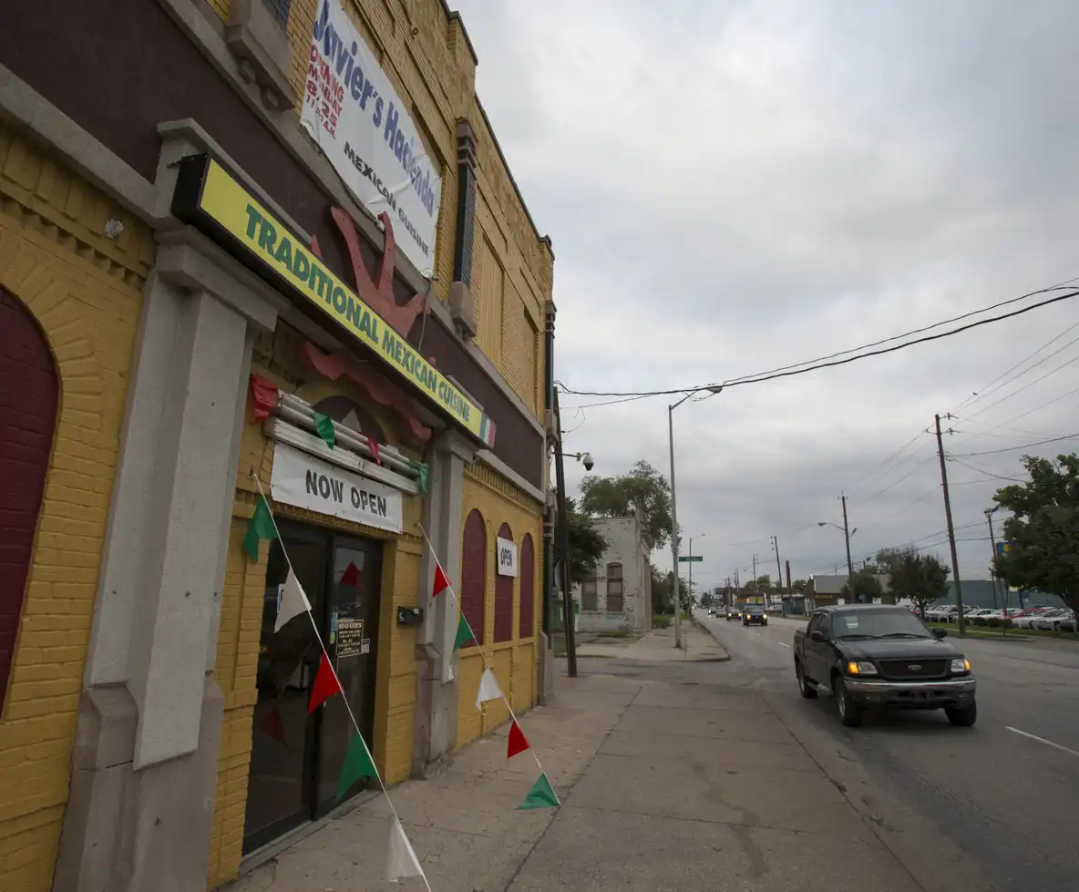 The storefront of a traditional-looking Mexican restaurant is shown. There is a "Now Open" sign over the front door and red, white and green pennants around the door.