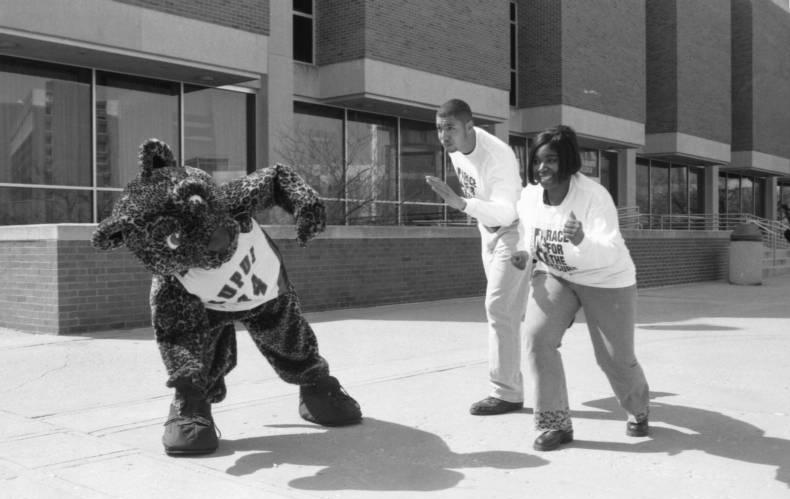 Two people and a Jaguar mascot are running outside of a building.
