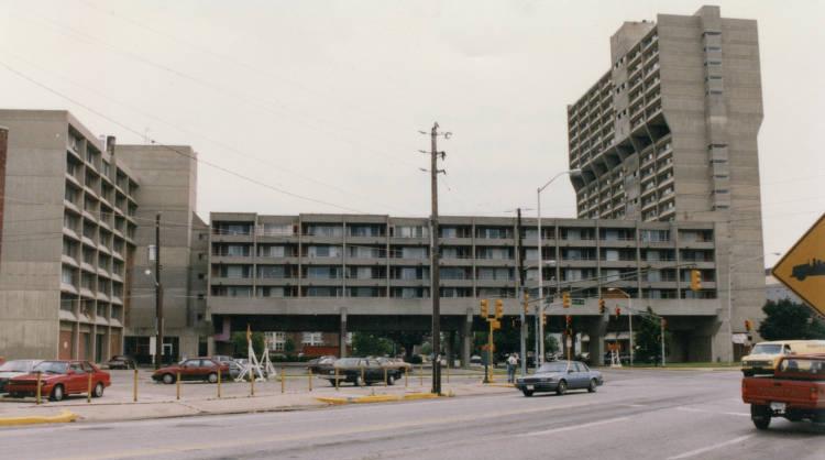 The front of a 1960's style stone multi-unit building. The center building is long, balconied and four-storied and raised aboveground by pylons. The wing on the right is a tower several stories higher. The left wing is much like the center building.