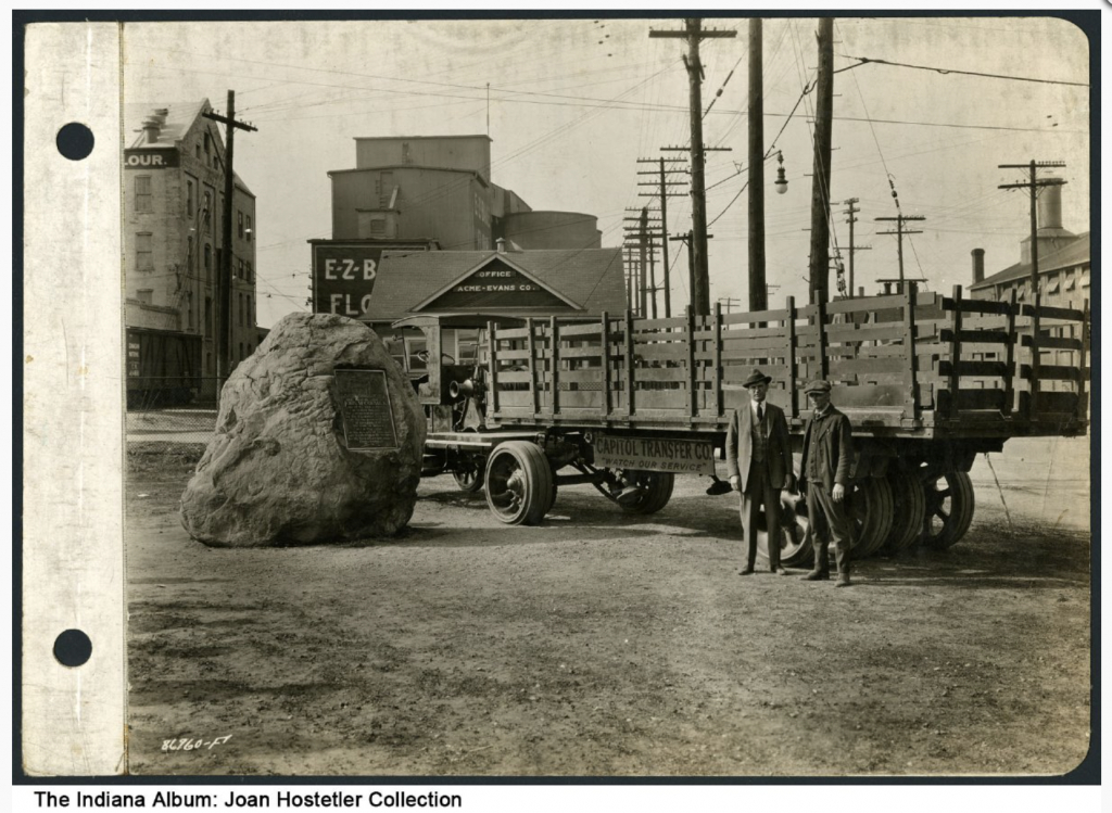 An old truck is parked next to a large boulder. Two men stand in front of the truck. 