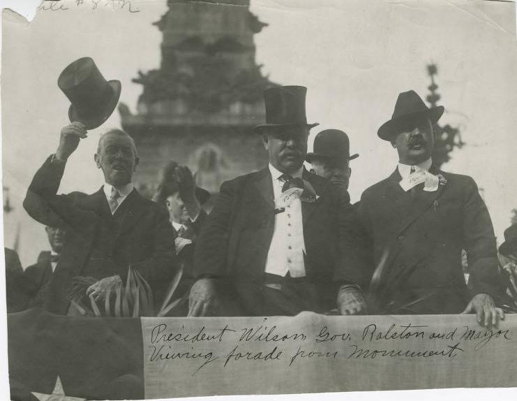 A group of men stand in front of the soldiers and sailors monument. A caption written on the photo reads: "President Wilson, Gov. Ralston and Mayor Viewing parade from monument