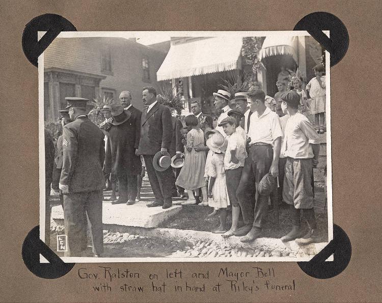 A group of people are standing. One man holds his hat over his heart. Written beneath the photo is "Gov. Ralston on left and Mayor Bell with straw hat in hand at Riley's funeral.