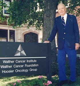 A man stands next to a sign that's in front of a building. The sign reads "Walther Cancer Institute, Walther Cancer Foundation, Hoosier Osteology Group."