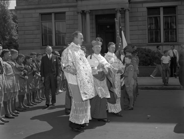 Several clergy men stand in the middle of a street. Boys in scout uniforms line up behind them. 