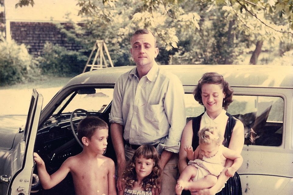 A family of five stand together in front of a car with the driver's side door open. 