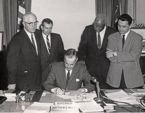 Four men stand behind a man seated at a desk signing a document.