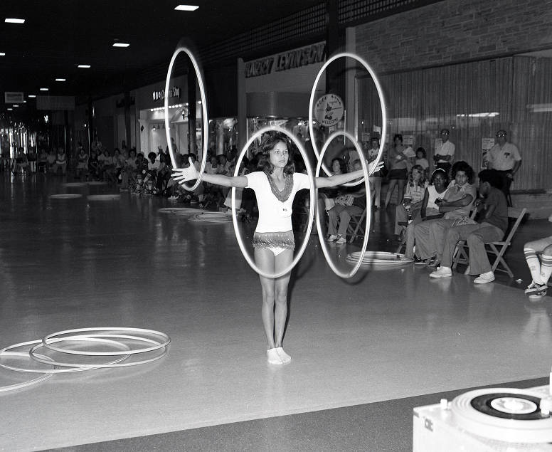 A woman performs in the middle of a shopping mall. She is holding up four hoola hoops