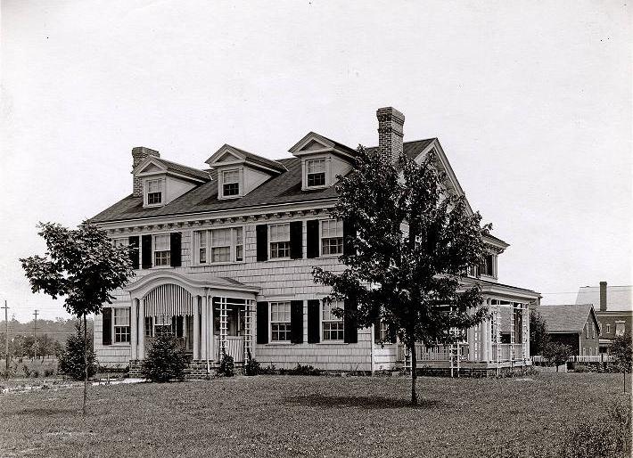 The house is a large, shingled, two-story, traditional with an arch-roofed portico at the entrance. It has dark shutters, a peaked roof with three gables and a chimney at each end. There is a covered side porch.