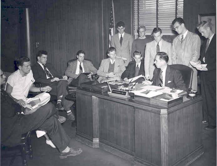 A group of men gather around a man seated at a desk.