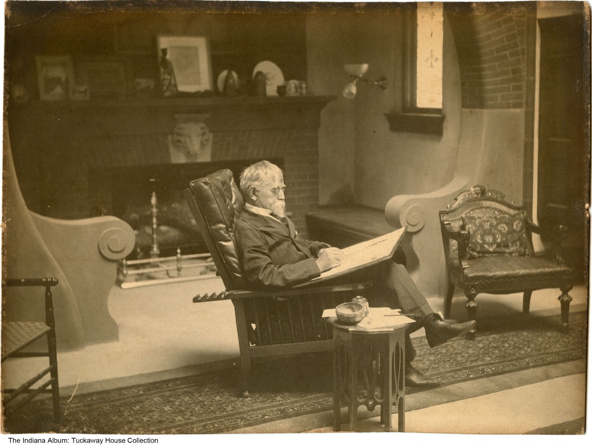 Lew Wallace is seated in the middle of a room with a writing board on his lap. The room has several chairs and a fireplace in the background.  