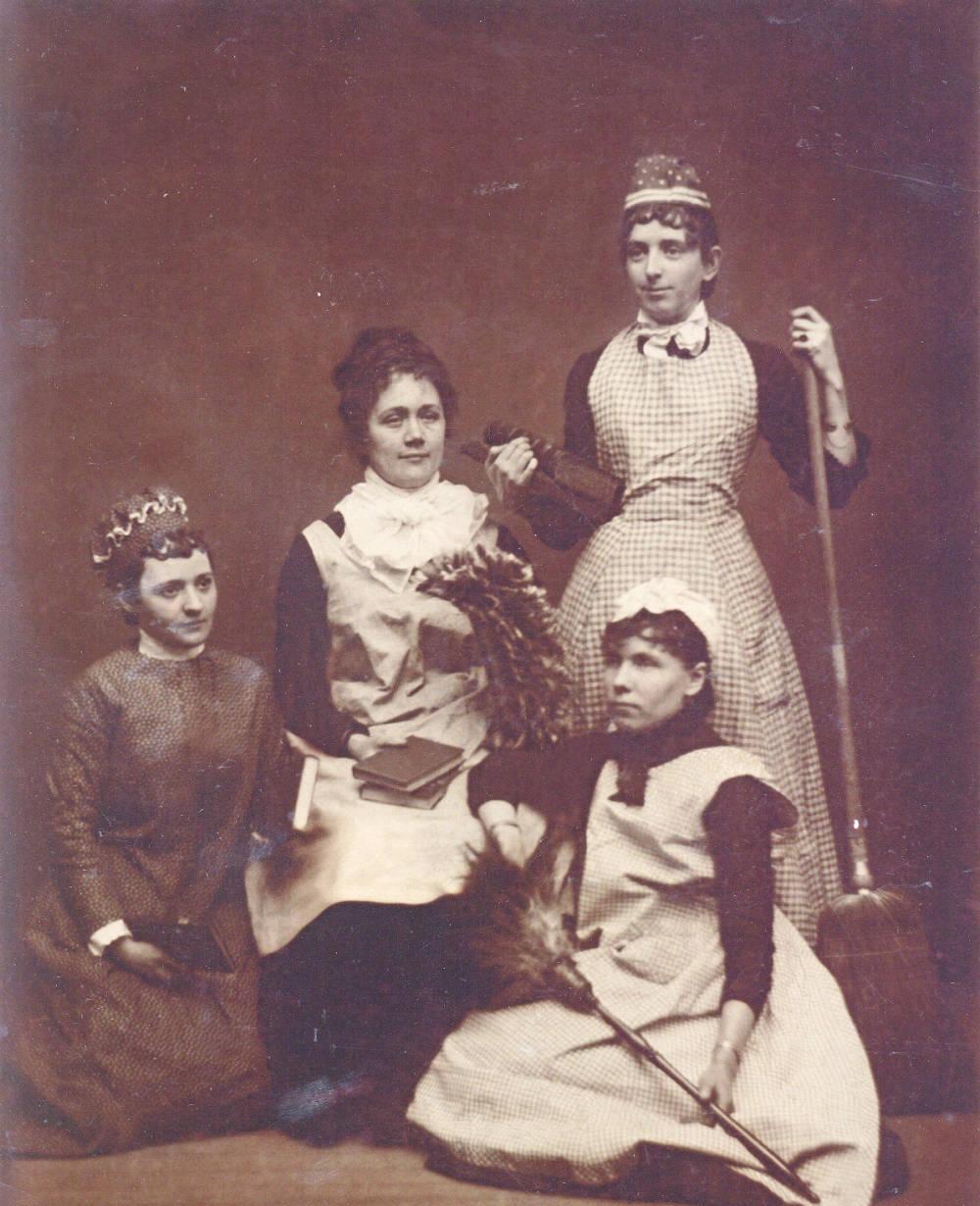 Four women are dressed in aprons and hold cleaning supplies.