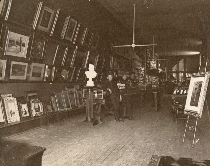 A couple of men lean on a glass case. The walls of the room are covered in framed images. 