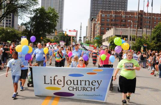 A group of people in a parade carry a banner with the Life Journey Church logo on it.