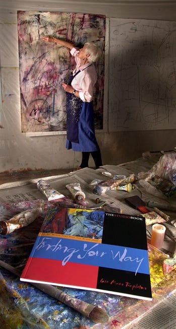 A woman stands and paints a canvas hung on a wall. 