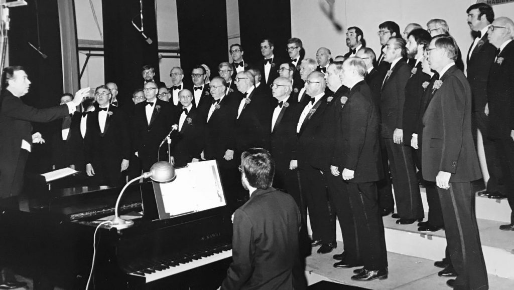 A choir of men stand together. A man is playing a piano.