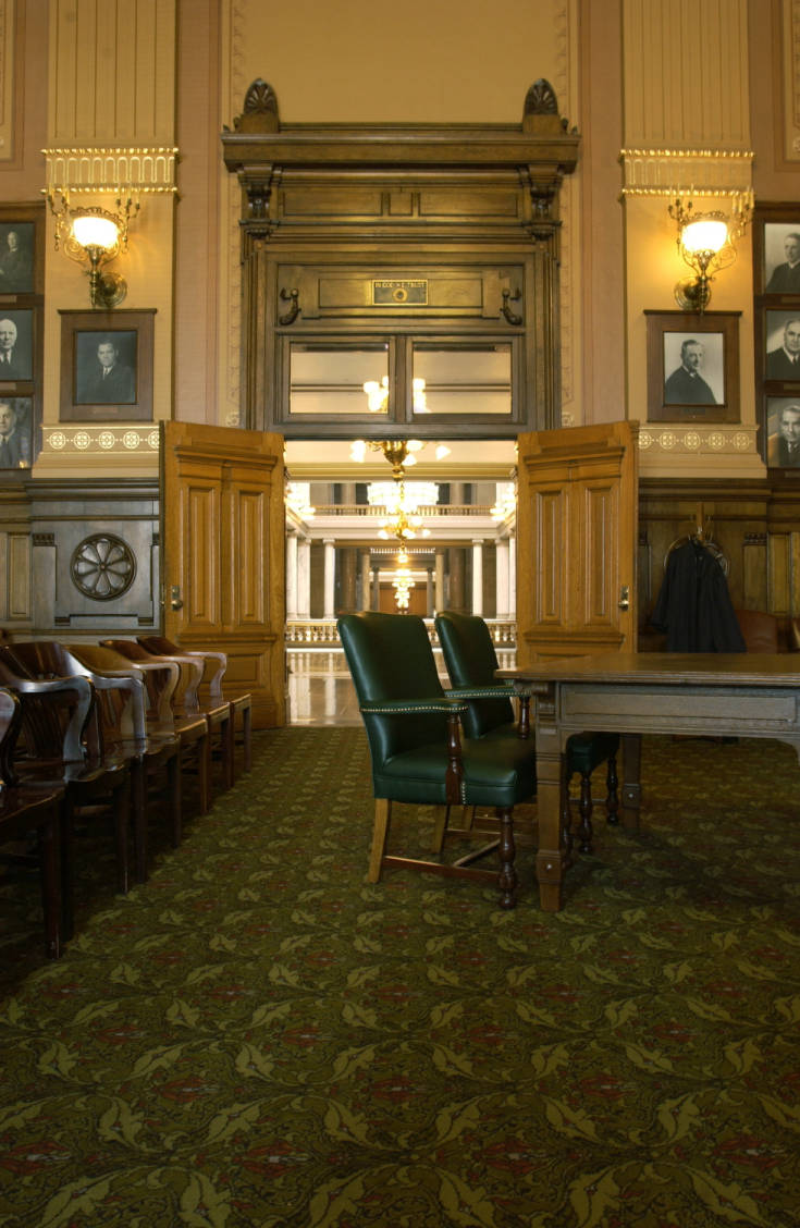 A grand, high-ceilinged room has jurors' chairs to the left and a wide, wooden table with two chairs to the right (for attorneys). Heavy wooden doors open onto a neoclassical foyer.