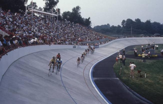 Several bicyclists race on a track with crowds of spectators watching from the stands.