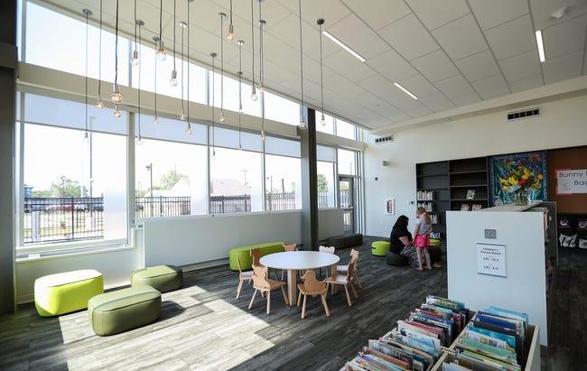 Interior view of a library space. Cushions and child-sized tables and chairs line the edge of the room by the windows. Shelves of books are set up on the other side.