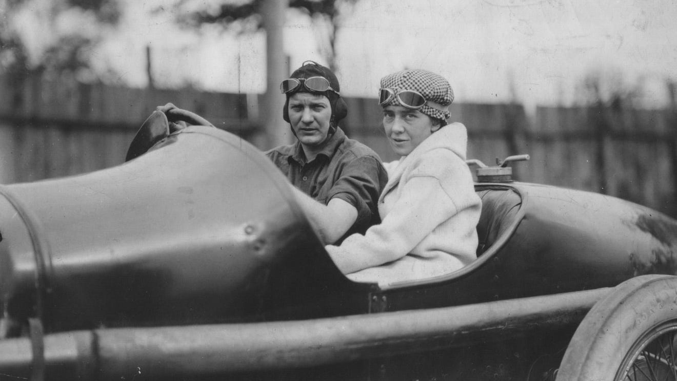 A man and a woman wearing motoring caps with goggles perched on their foreheads, sit together in the open seat of a race car.