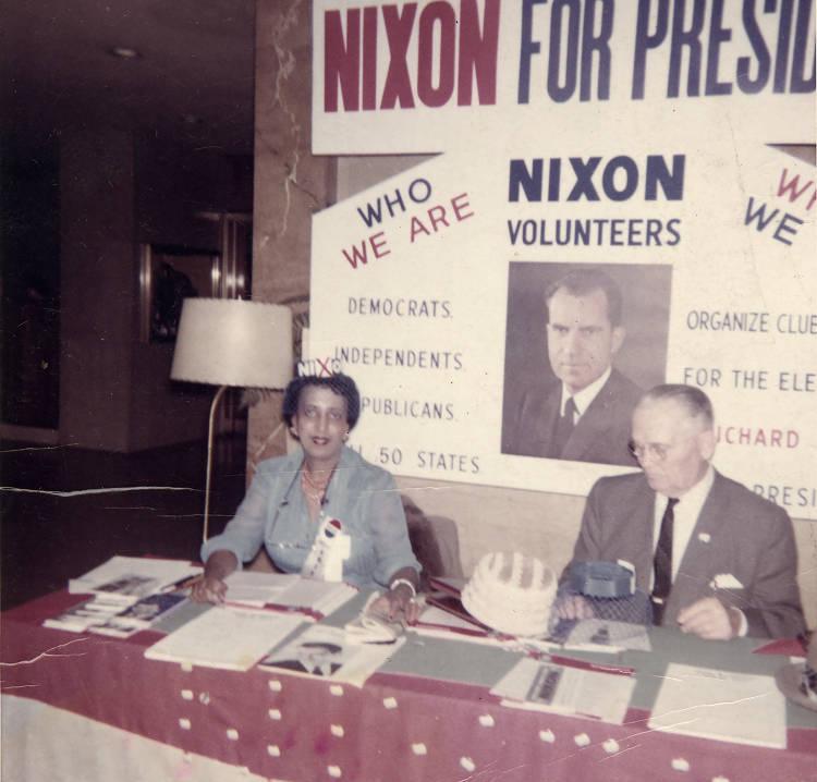 A man and a woman sit at a table that is covered with pamphlets and flyers. Behind them is a "Nixon For President" sign with a picture of Richard Nixon on it.