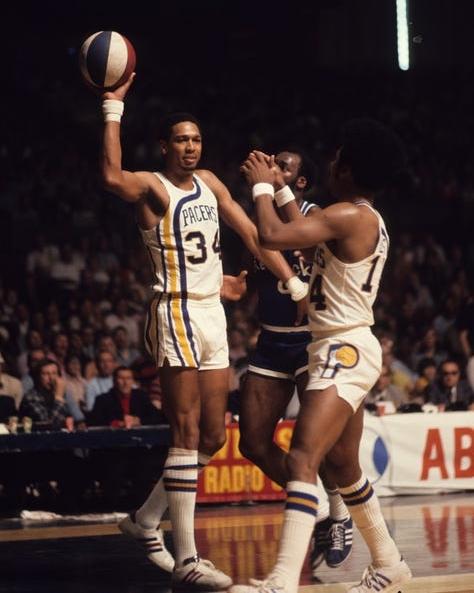 There are three men in basketball uniforms. One of them, Daniels, holds a ball up preparing to pass.