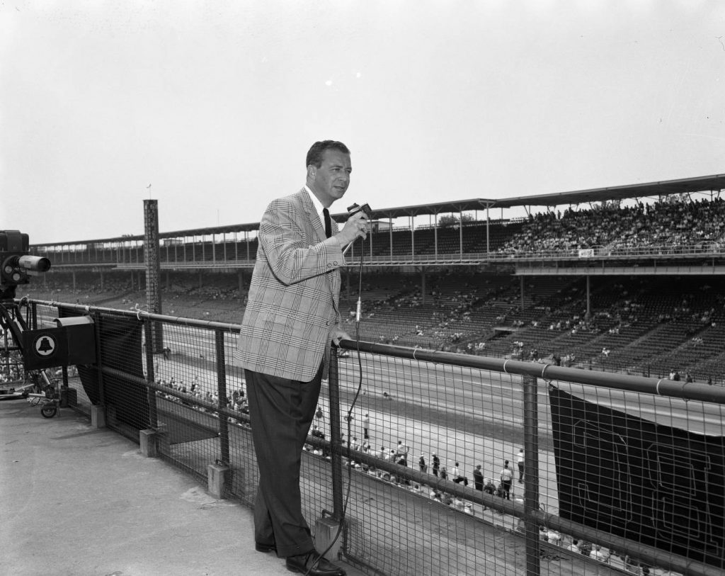 Sid Collins stands along fencing and speaks into a microphone. He is looking towards the race track in the background.