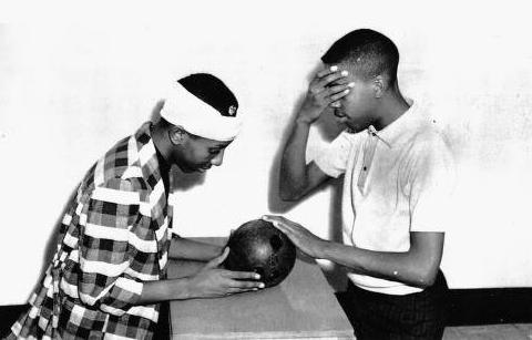 Two boys enact a scene from a play. One boy holds a ball on a table while the other covers his eyes with one hand and touches the ball with the other.