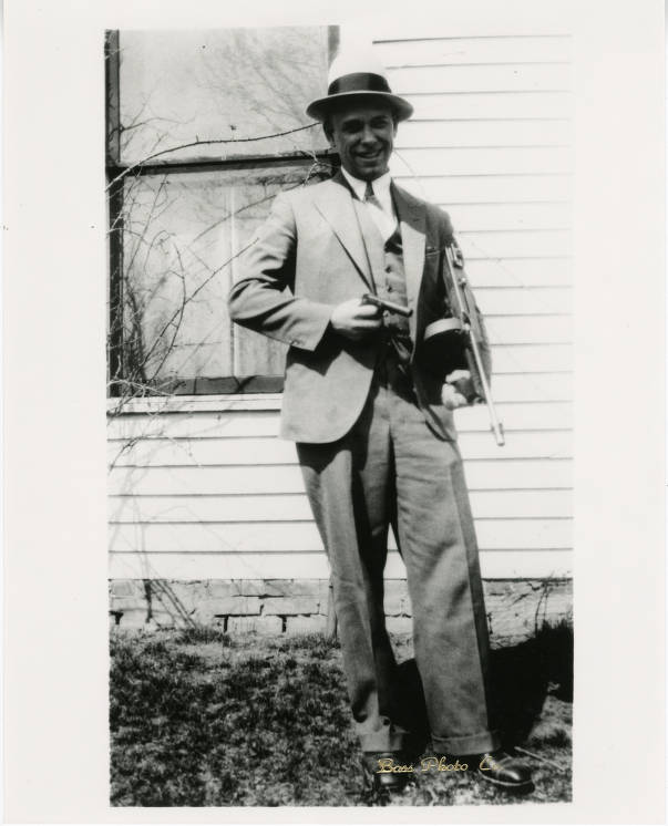 A man wearing a three-piece suit and a hat is holding a handgun and a tommy (machine) gun.